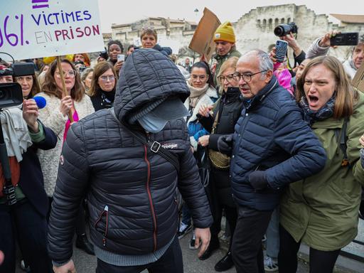 Ein maskierter Angeklagter im Vergewaltigungsprozess von Avignon geht zum Gericht. Um ihn herum sind wütende Menschen zu sehen.