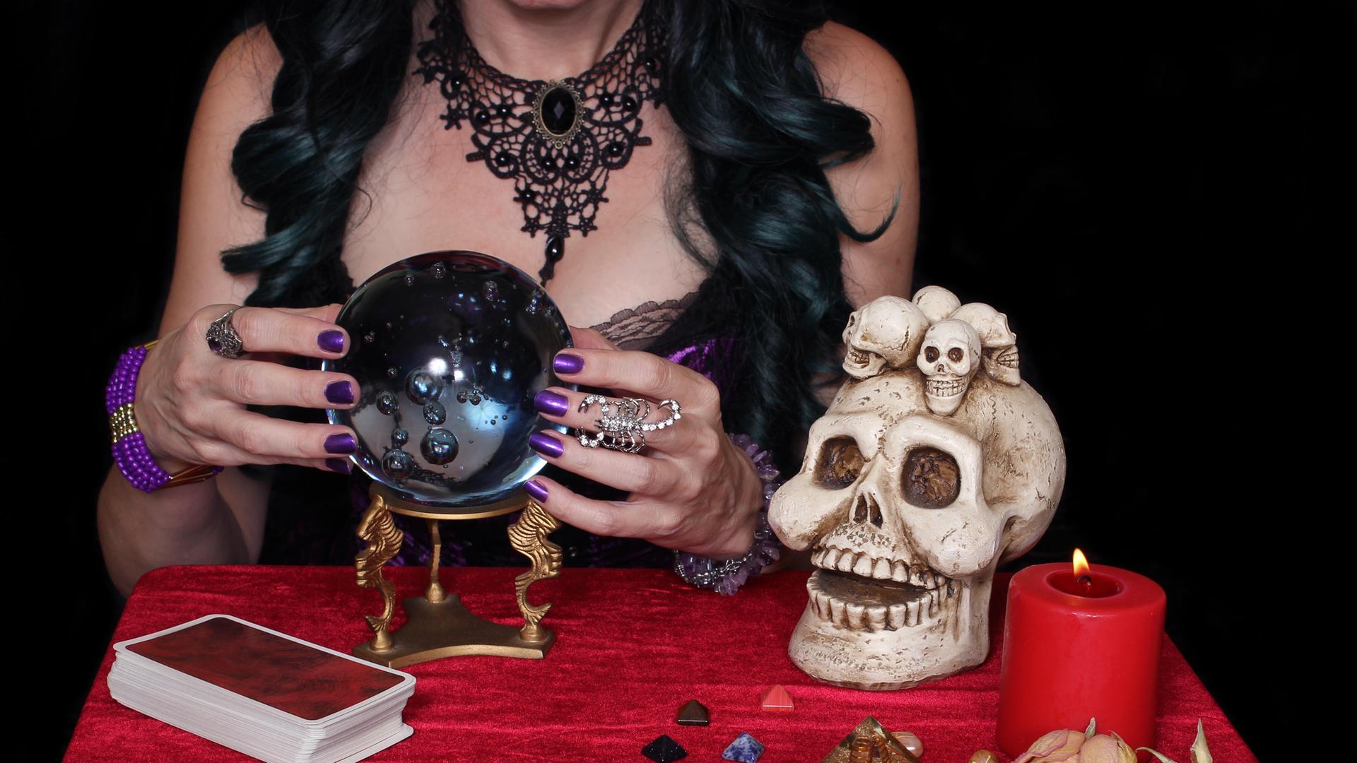 Eine Frau mit langen schwarzen Haaren sitzt an einem Tisch, auf dem ein Totenkopf, eine Kerze und Tarot-Karten liegen. Die Frau umfasst eine Kristallkugel.