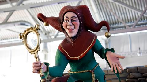 Die Figur eines Narren mit Spiegel und Narrenkappe zwinkert und streckt die Zunge heraus. 