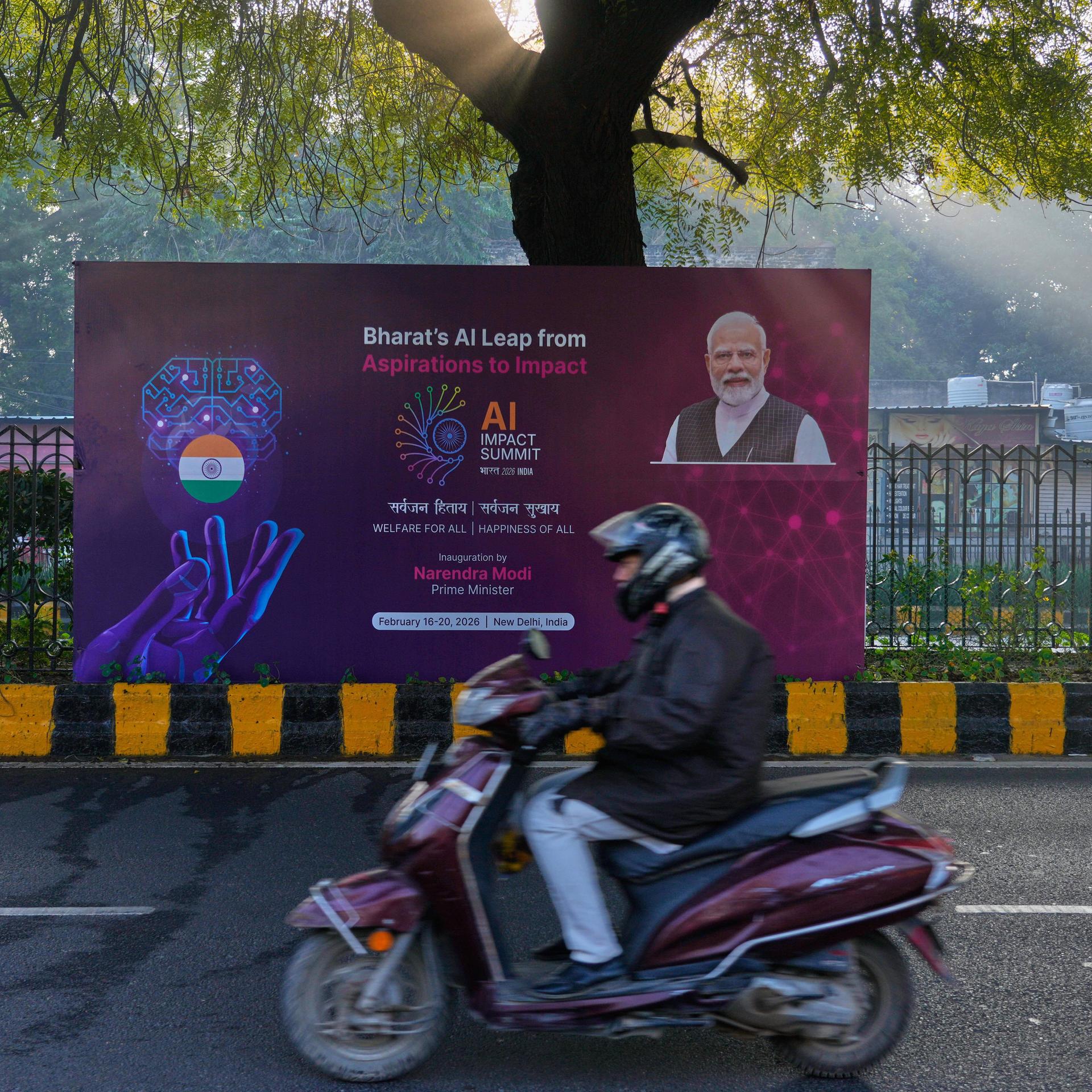 Neu Delhi: Ein Rollerfahrer fährt an einem Banner des KI-Gipfels mit dem indischen Premierminister Narendra Modi vorbei, das auf einer Straße zum Veranstaltungsort des Gipfels steht.