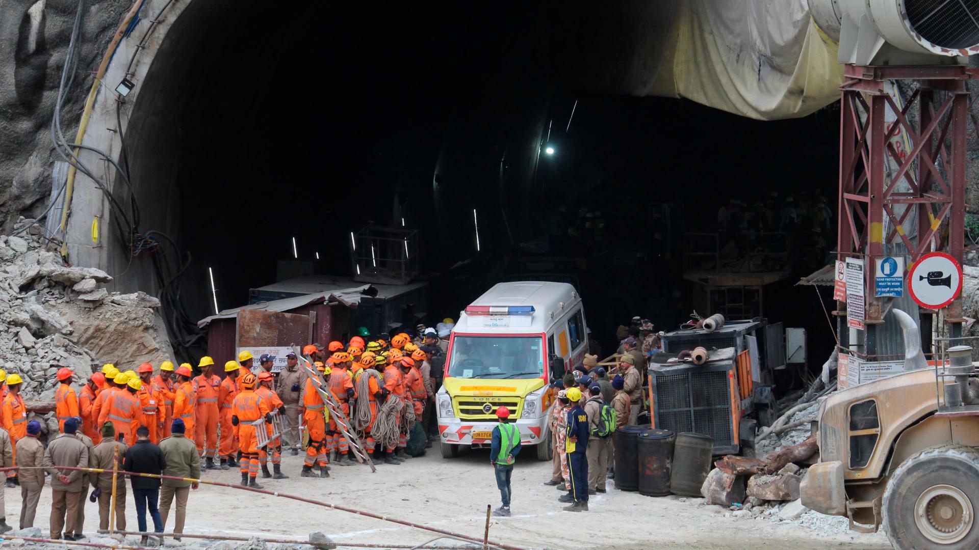 Vor dem riesigen Tunnel stehen viele Rettungs-Kräfte in orangefarbenen Anzügen und das Rettungs-Fahrzeug.