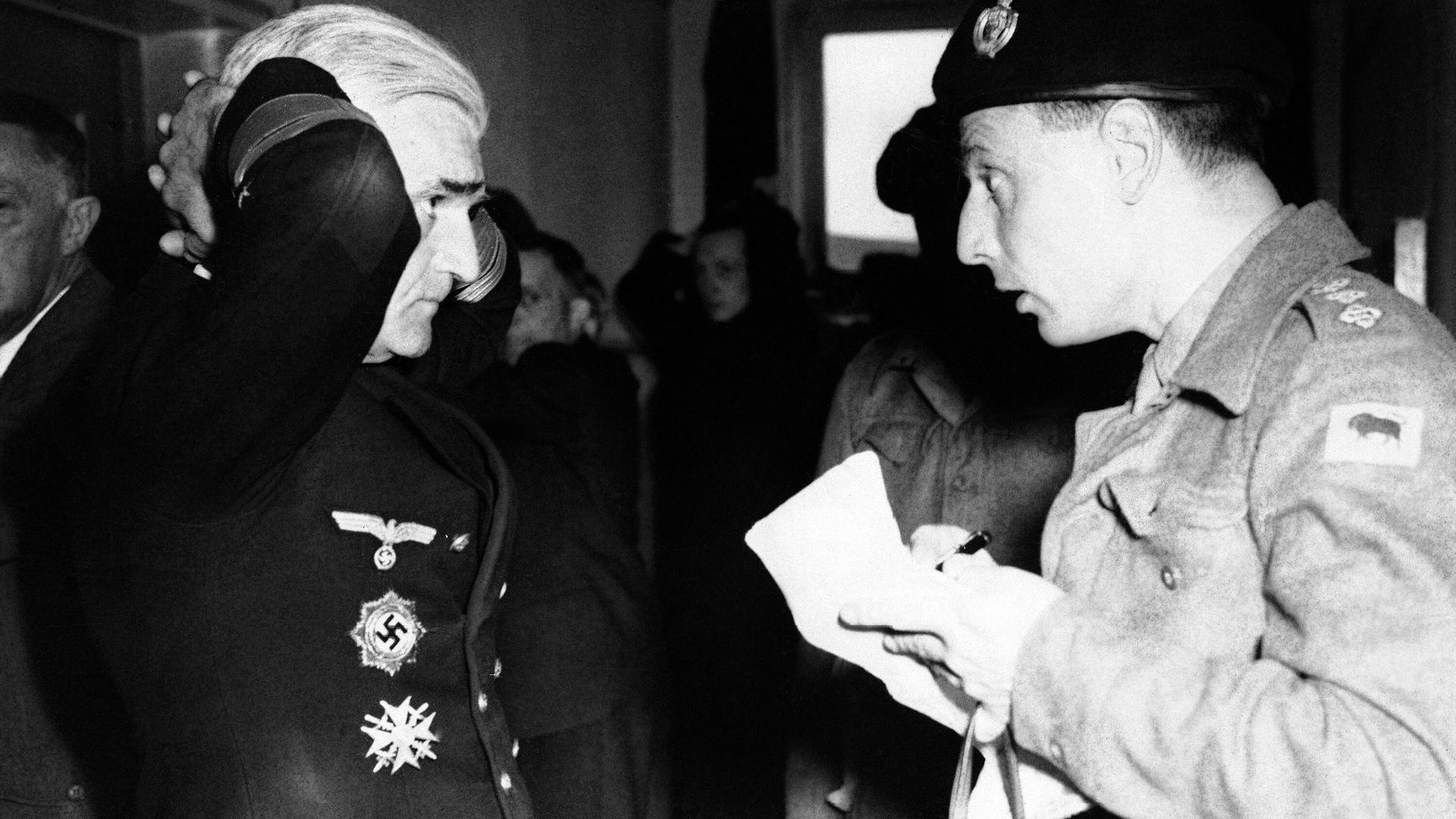 Nazi-Admiral Gerhard Wagner steht mit hinter dem Kopf verschränkten Händen vor einem britischen Soldaten und beantwortet Fragen.