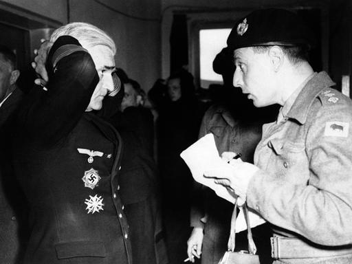 Nazi-Admiral Gerhard Wagner steht mit hinter dem Kopf verschränkten Händen vor einem britischen Soldaten und beantwortet Fragen.