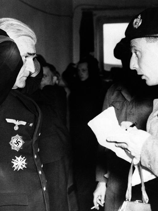 Nazi-Admiral Gerhard Wagner steht mit hinter dem Kopf verschränkten Händen vor einem britischen Soldaten und beantwortet Fragen.