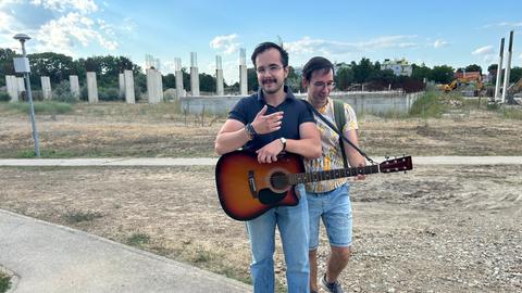 Zwei Männer, einer mit Gitarre, stehen vor einer Bauruine im kroatischen Vukovar