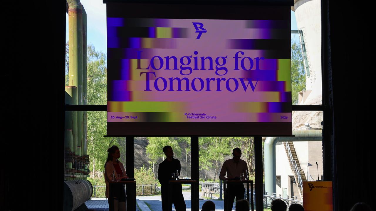 Die Organisatoren stellen das Programm der Ruhrtriennale unter dem Motto "Longing for Tomorrow" vor. Zu sehen ist eine Industriehalle, in der mehrere Personen vor Rednerpulten stehen; im Hintergrund ein großes Plakat mit dem Motto des Festivals.