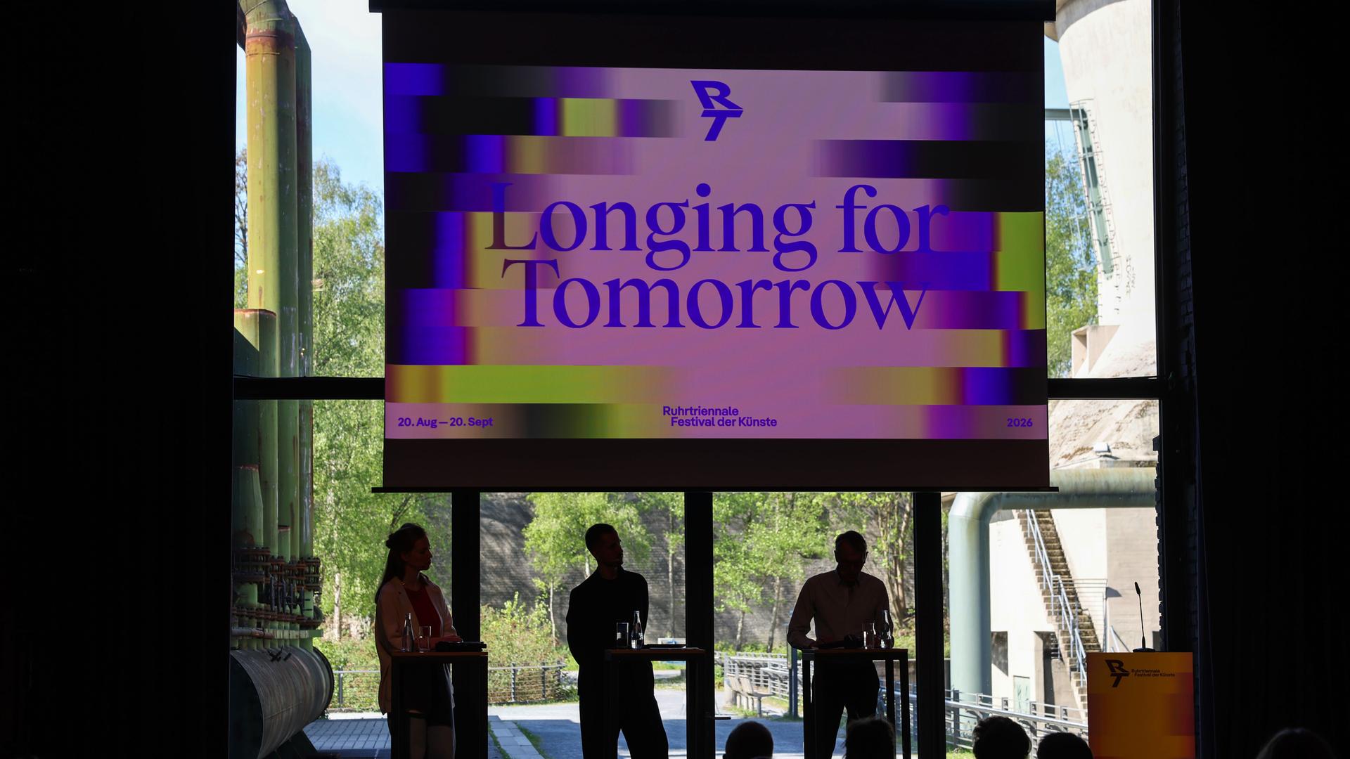 Die Organisatoren stellen das Programm der Ruhrtriennale unter dem Motto "Longing for Tomorrow" vor. Zu sehen ist eine Industriehalle, in der mehrere Personen vor Rednerpulten stehen; im Hintergrund ein großes Plakat mit dem Motto des Festivals.