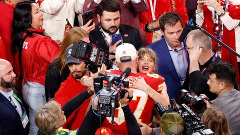 Taylor Swift und Travis Kelce umarmen sich nach dem Super Bowl. Mehrere Kameras sind auf sie gerichtet.