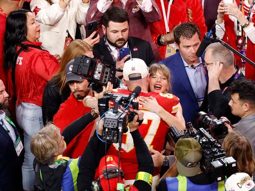 Taylor Swift und Travis Kelce umarmen sich nach dem Super Bowl. Mehrere Kameras sind auf sie gerichtet.