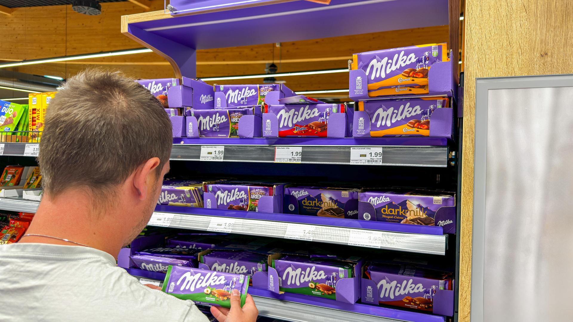 Ein Mann nimmt im Supermarkt eine Tafel Milka-Schokolade aus dem Regal. Er schaut auf die Packung. Der Mann ist schräg von hinten zu sehen. Ein Mann nimmt im Supermarkt eine Tafel Milka-Schokolade aus dem Regal. Er schaut auf die Packung. Der Mann ist schräg von hinten zu sehen.