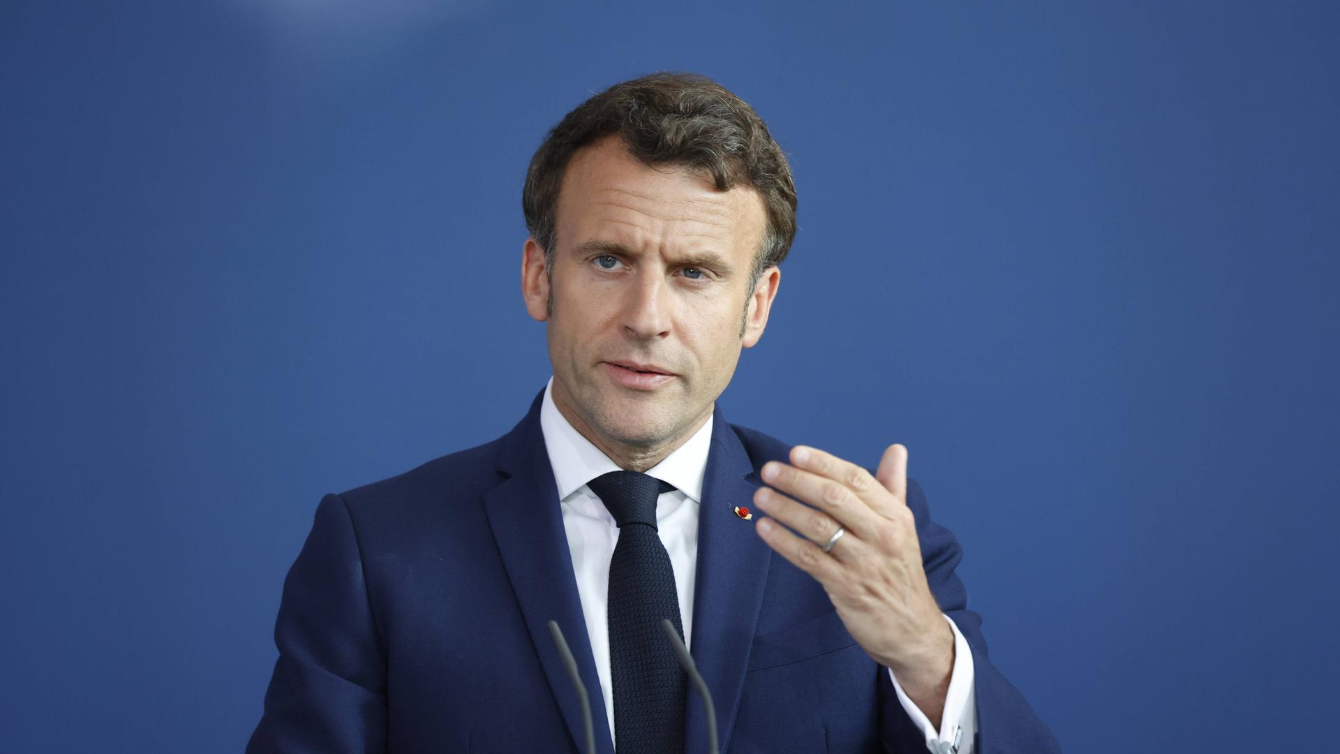 Macron gestikuliert bei einer Pressekonferenz mit einer Hand. Er schaut in Richtung der Kamera. 