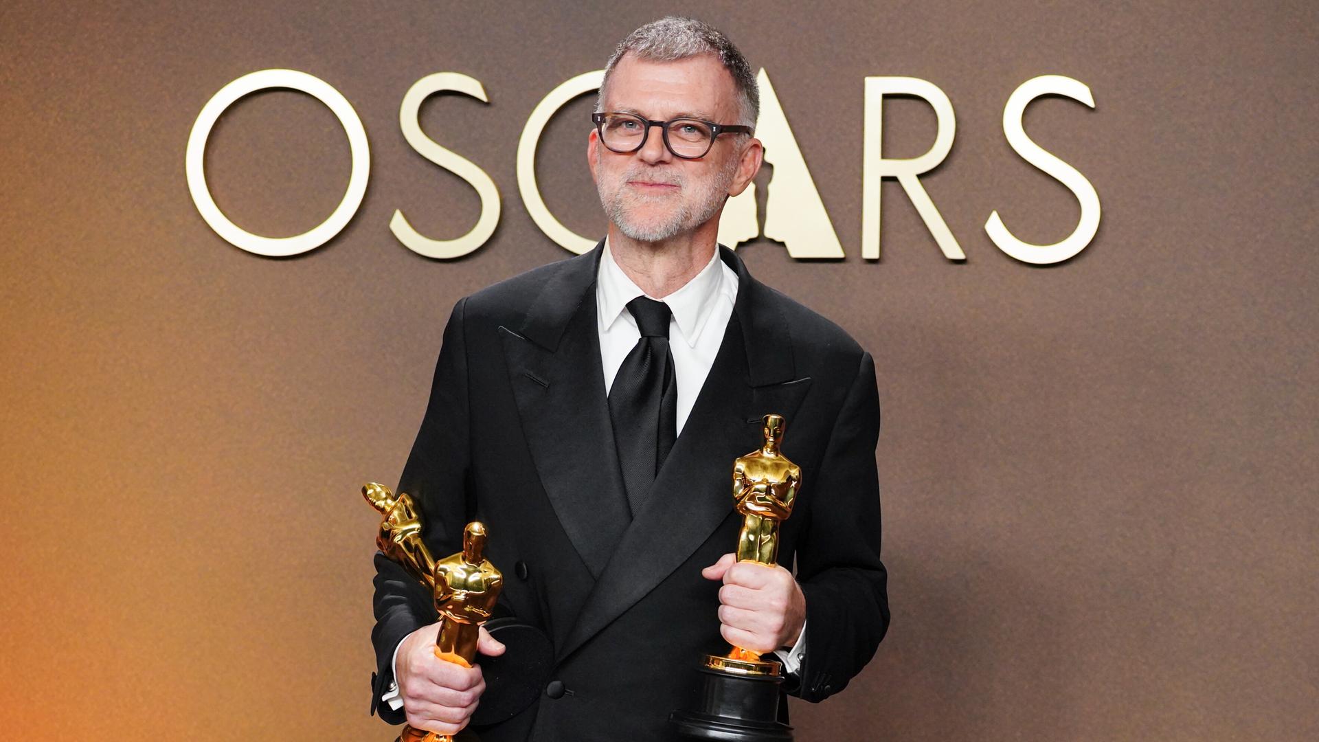 Das ist der Regisseur Paul Thomas Anderson. Er steht im Anzug auf der Bühne. Er hat in jeder Hand einen Oscar. Das ist der Regisseur Paul Thomas Anderson. Er steht im Anzug auf der Bühne. Er hat in jeder Hand einen Oscar.