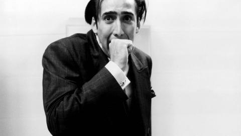 Im Still aus "Vampire's Kiss" beißt Nicolas Cage sich mit gequältem Gesichtsausdruck in die eigene Hand.