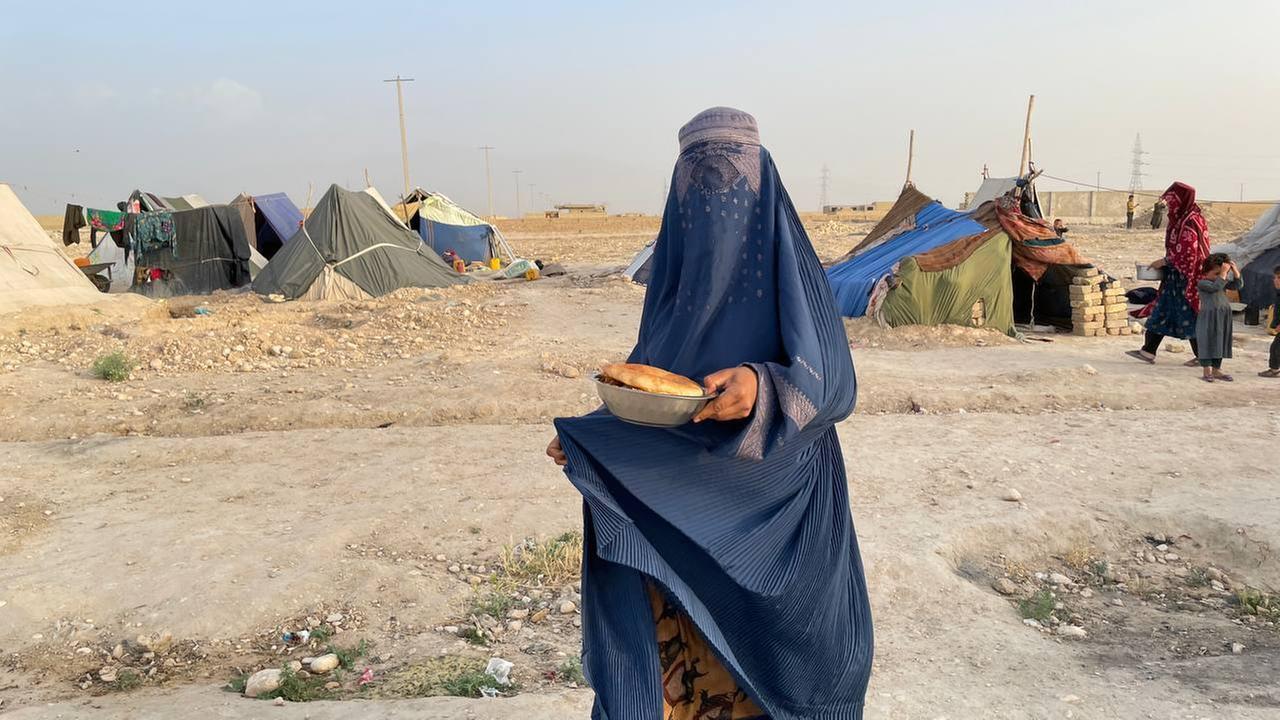 Eine Frau trägt eine blaue Burka, die auch ihr Gesicht verhüllt, in der Hand hält sie eine Schüssel mit Brot. Im Hintergrund: Zelte in einer staubigen Landschaft. Eine Frau trägt eine blaue Burka, die auch ihr Gesicht verhüllt, in der Hand hält sie eine Schüssel mit Brot. Im Hintergrund: Zelte in einer staubigen Landschaft.