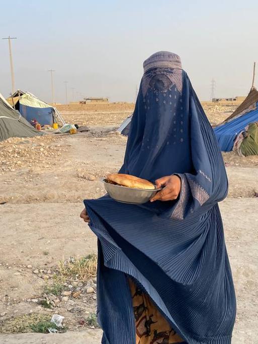 Eine Frau trägt eine blaue Burka, die auch ihr Gesicht verhüllt, in der Hand hält sie eine Schüssel mit Brot. Im Hintergrund: Zelte in einer staubigen Landschaft.