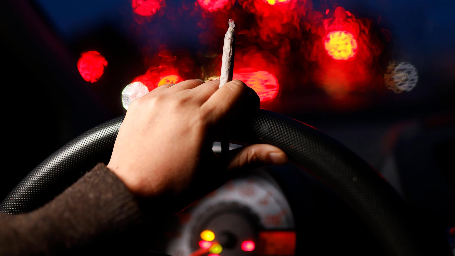 Aus der Perspektive eines Autofahrers sind seine Hände am Lenkrad bei einer Nachtfahrt zu sehen. In einer Hand hält er einen Cannabis-Joint während verschwommene Lichter hinter der Frontscheibe zu sehen sind.