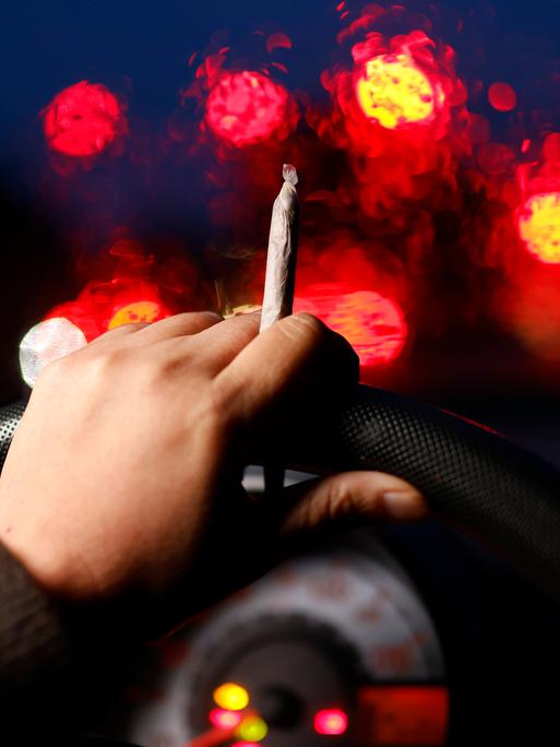 Aus der Perspektive eines Autofahrers sind seine Hände am Lenkrad bei einer Nachtfahrt zu sehen. In einer Hand hält er einen Cannabis-Joint während verschwommene Lichter hinter der Frontscheibe zu sehen sind.