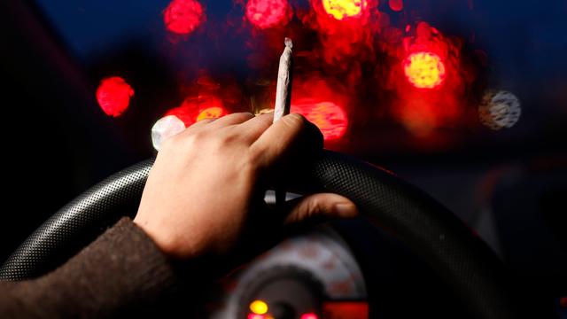 Aus der Perspektive eines Autofahrers sind seine Hände am Lenkrad bei einer Nachtfahrt zu sehen. In einer Hand hält er einen Cannabis-Joint während verschwommene Lichter hinter der Frontscheibe zu sehen sind.