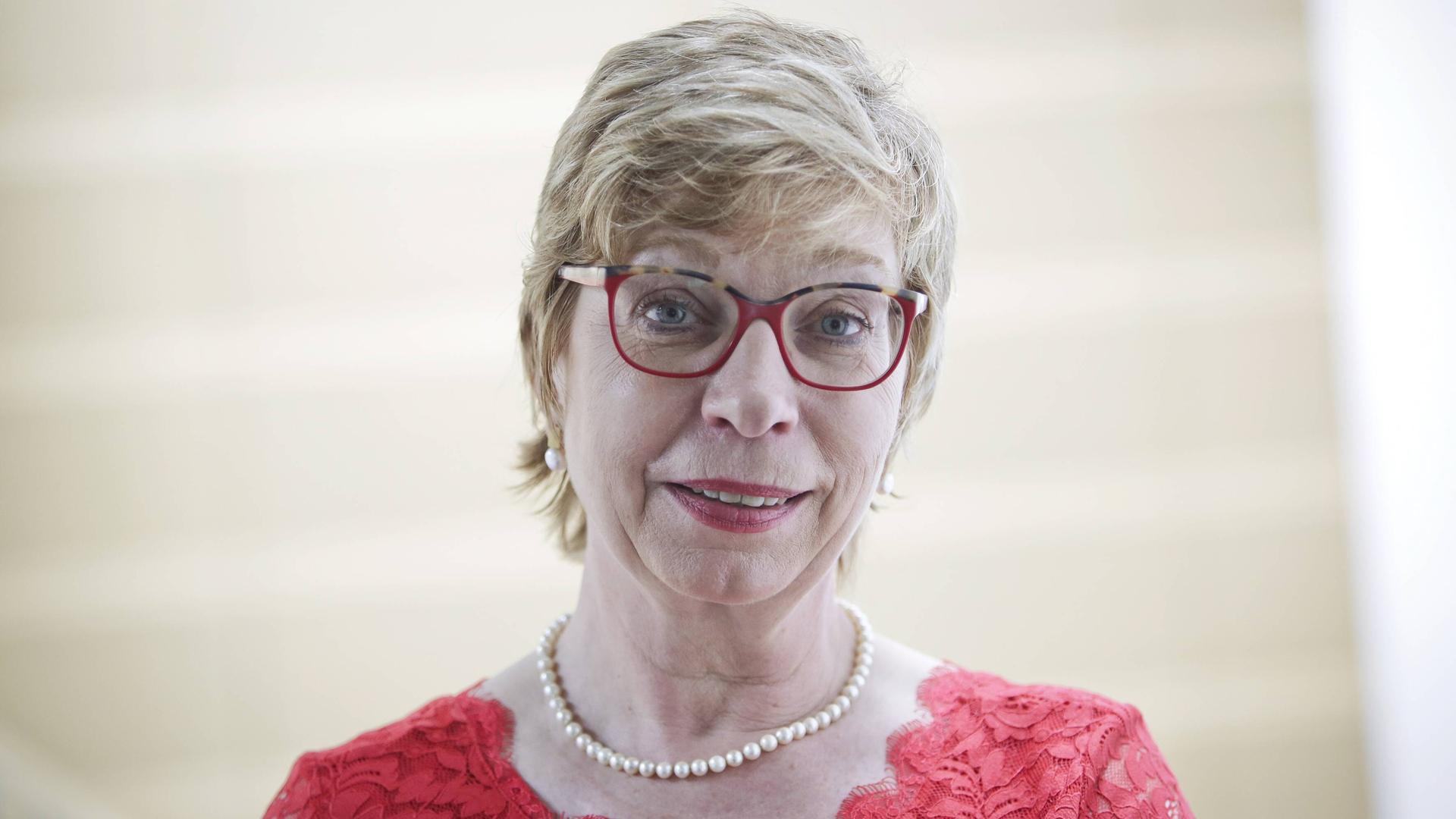 Ein Porträt von Susanne Lin-Klitzing, Bundesvorsitzende des Deutschen Philologenverbandes, mit Perlenkette, roter Brille und rotem-spitzen Kleid vor einem hellem Hintergrund.