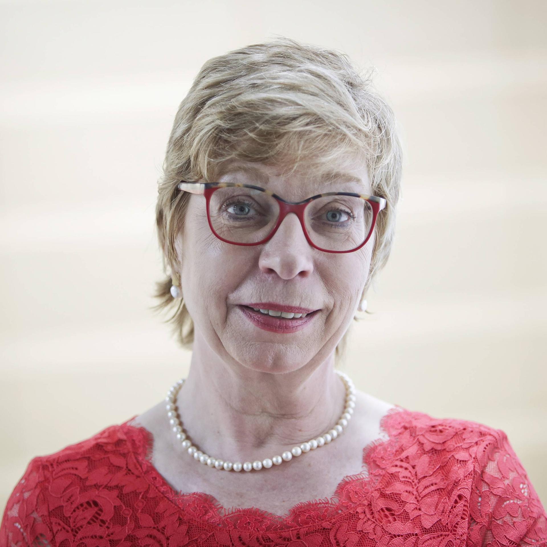 Porträt von Prof. Dr. Susanne Lin-Klitzing, Professorin für Schulpädagogik und Bundesvorsitzende des Deutschen Philologenverbandes mit Perlenkette, roter Brille und rotem Kleid vor hellem Hintergrund.