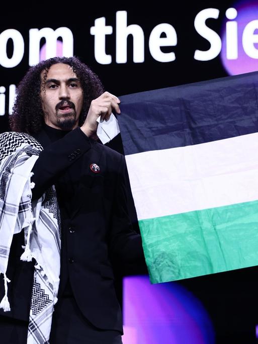 Abdallah Alkhatib, Regisseur vom Film "Chronicles from the Siege" spricht während der Preisverleihung bei der Abschlussgala im Berlinale Palast auf der Bühne. Neben ihm wird die palästinensische Fahne hochgehalten.