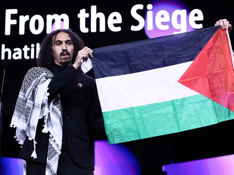 Abdallah Alkhatib, Regisseur vom Film "Chronicles from the Siege" spricht während der Preisverleihung bei der Abschlussgala im Berlinale Palast auf der Bühne. Neben ihm wird die palästinensische Fahne hochgehalten.