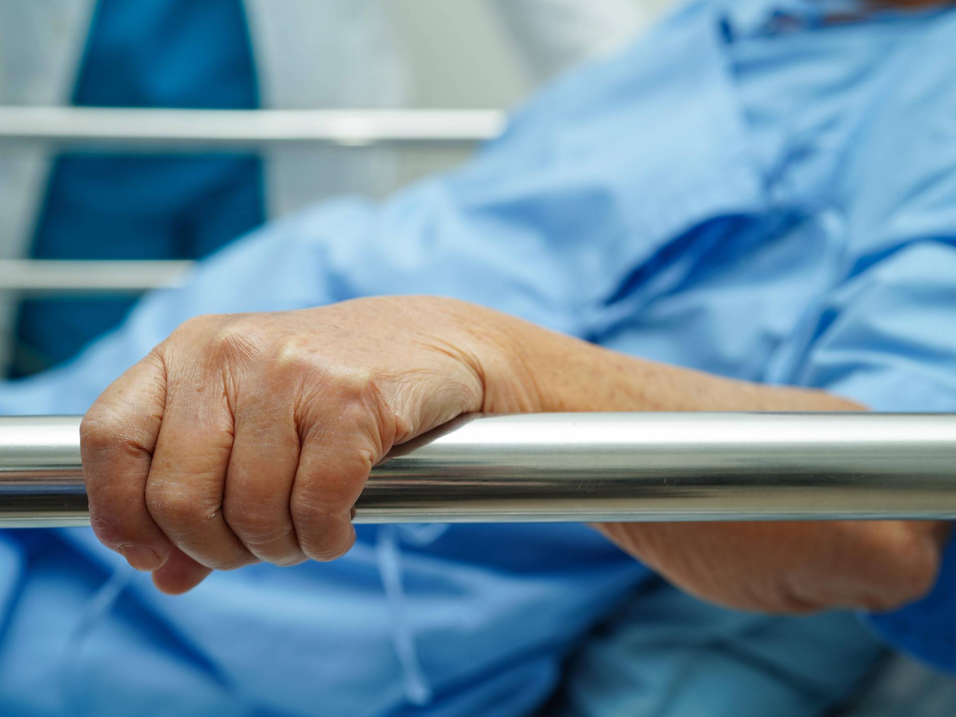 Eine ältere Frau trägt einen Krankenhauskittel und hält sich an der Seite ihres Krankenhausbettes fest. 
