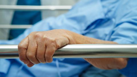 Eine ältere Frau trägt einen Krankenhauskittel und hält sich an der Seite ihres Krankenhausbettes fest. 
