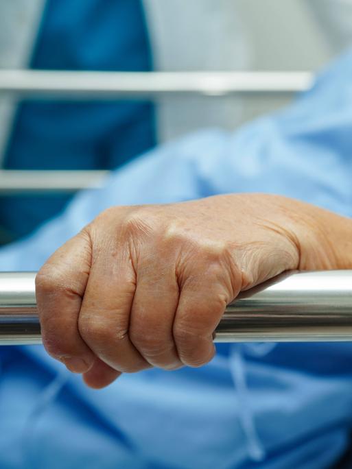 Eine ältere Frau trägt einen Krankenhauskittel und hält sich an der Seite ihres Krankenhausbettes fest. 