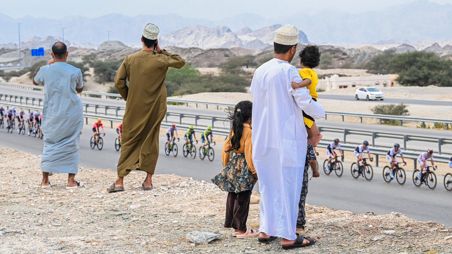 Tour Of Oman 2024. Zu sehen sind Zuschauer in traditioneller Kleidung von hinten während der 2. Etappe der 13. Ausgabe der Tour of Oman. Im Hintergrund karge Wüstenlandschaft und eine Autobahn mit dem Peloton.