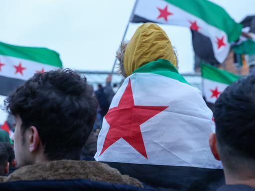 Ein Kind, was nur von hinten zu sehen ist, sitzt auf den Schultern eines Erwachsenen. Es trägt die syrische Flagge um die Schultern. Sie stehen einer Menschenmenge in der weitere Menschen mit syrischer Flagge von hinten zu sehen sind.