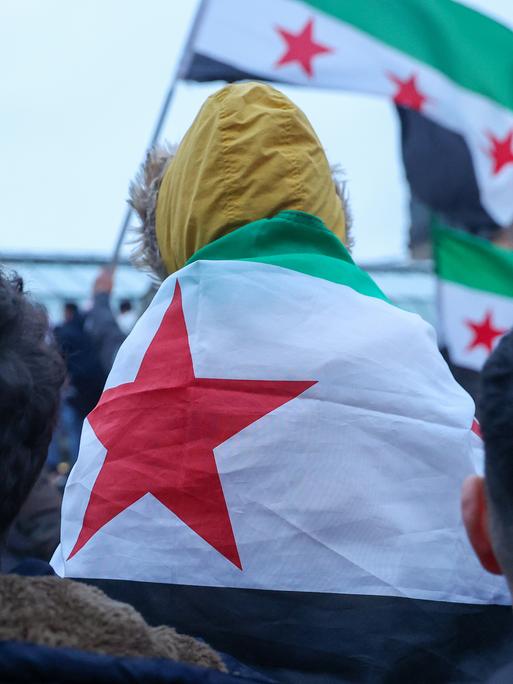 Ein Kind, was nur von hinten zu sehen ist, sitzt auf den Schultern eines Erwachsenen. Es trägt die syrische Flagge um die Schultern. Sie stehen einer Menschenmenge in der weitere Menschen mit syrischer Flagge von hinten zu sehen sind.