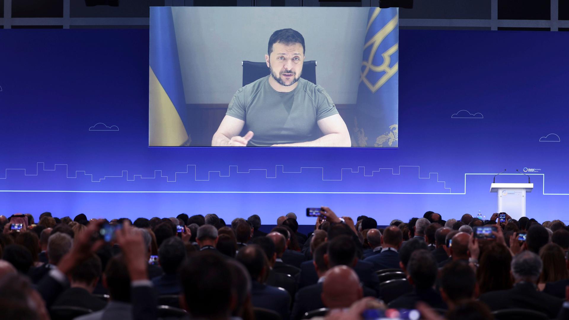 Wolodymyr Selenskyj ist auf einer Video-Leinwand zu sehen. Davor sitzen viele Teilnehmerinnen und Teilnehmer von der Geber-Konferenz. 