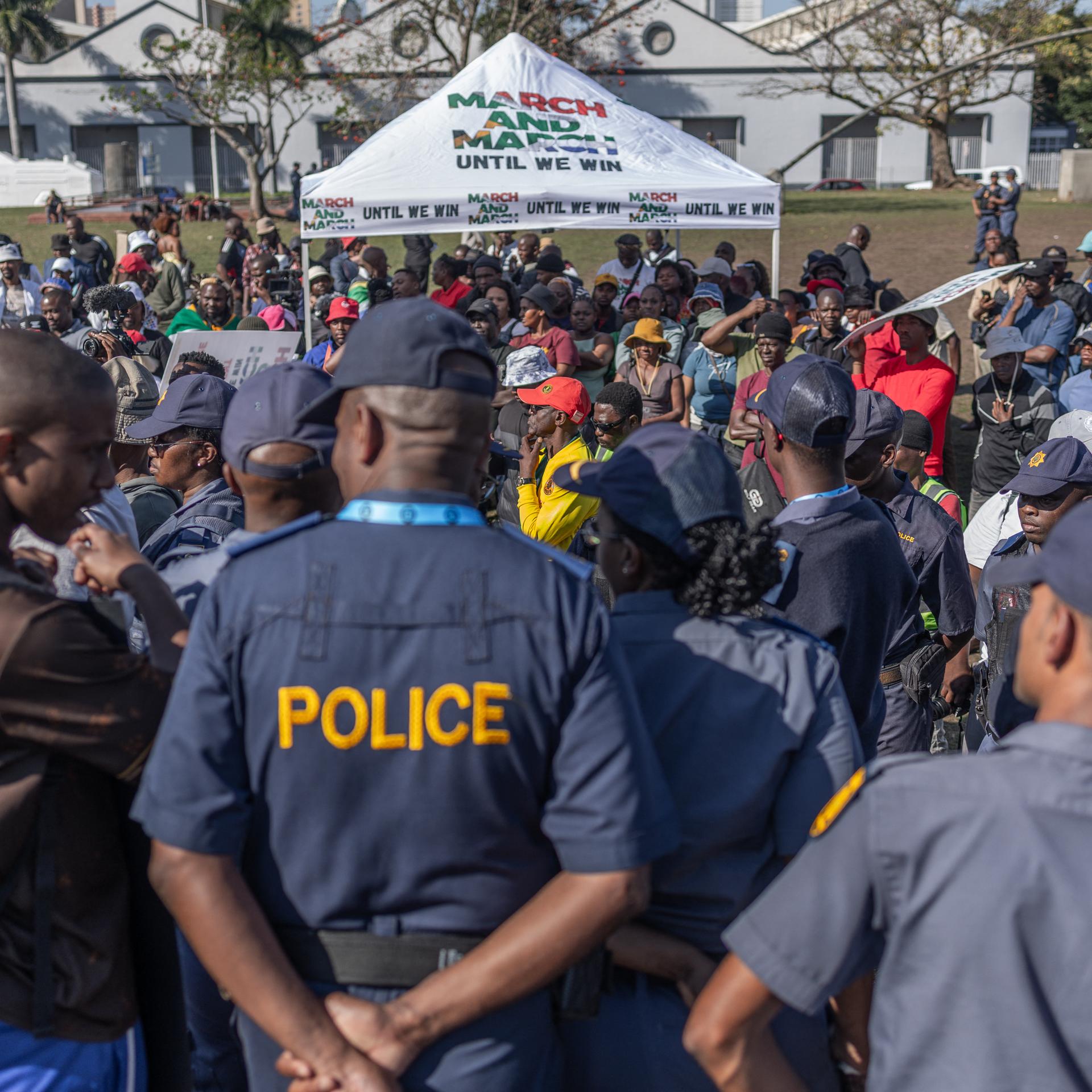 Vor dem Konferenzzentrum des SADC in Durban stehen Menschen und protestieren, im Vordergrund stehen zwei Polizisten.