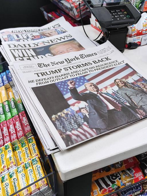 Auf einem Verkaufstisch eines Kiosks liegen verschiedene Zeitungen aus. Auf den Titelseiten geht es um den Sieg von Donald Trump bei der Präsidentschaftswahl 2024.