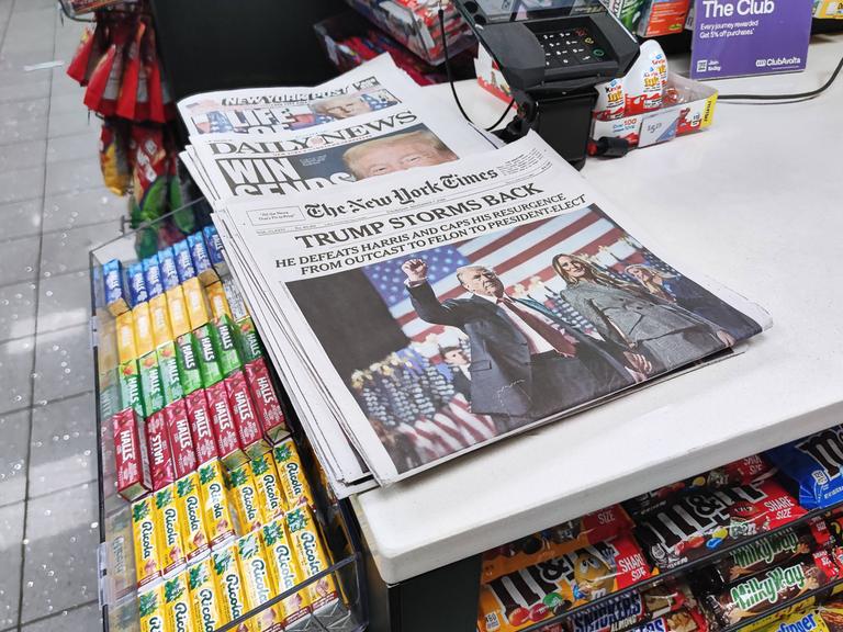 Auf einem Verkaufstisch eines Kiosks liegen verschiedene Zeitungen aus. Auf den Titelseiten geht es um den Sieg von Donald Trump bei der Präsidentschaftswahl 2024.