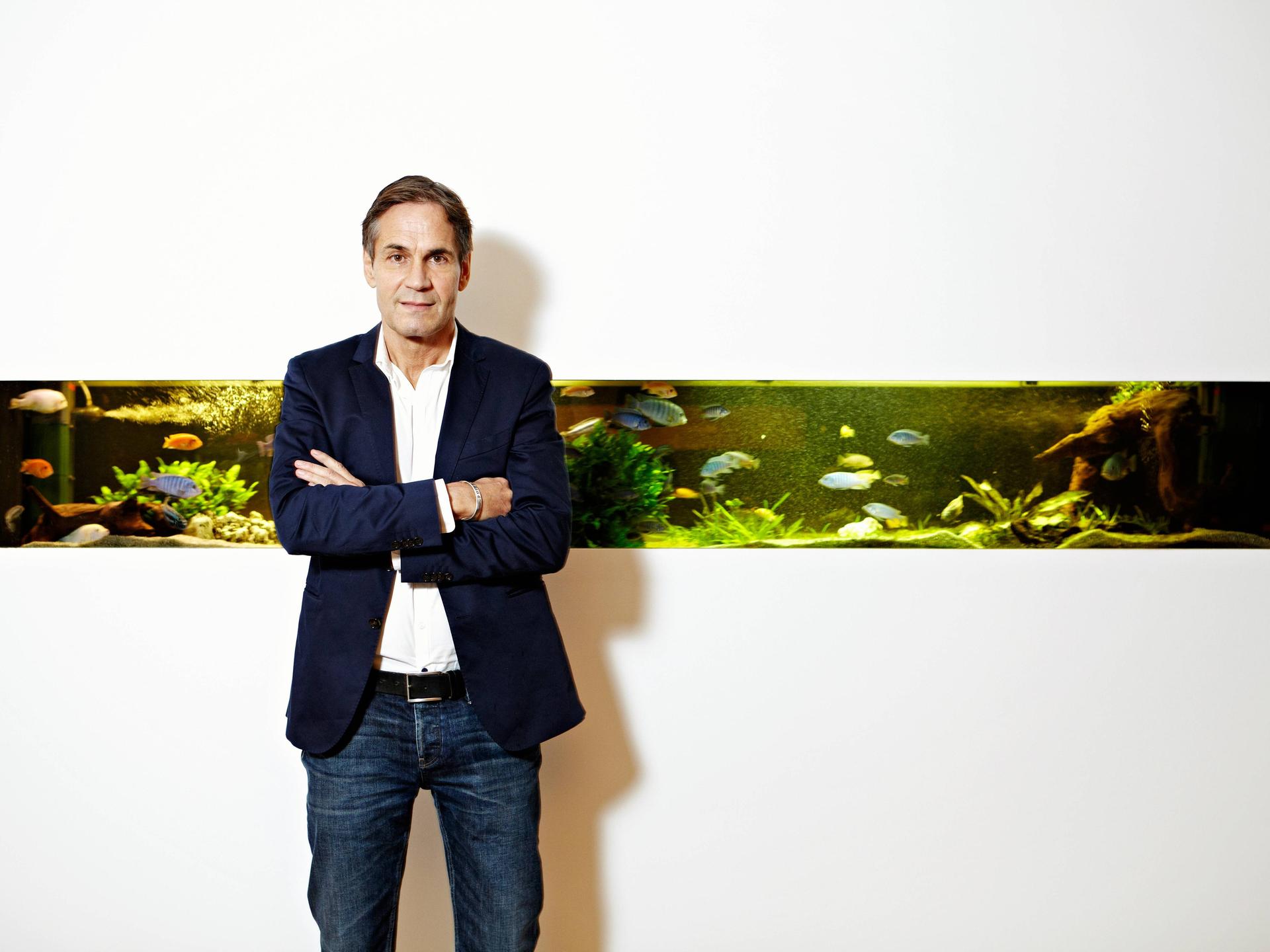 Jean Remy von Matt vor dem Aquarium der Werbeagentur Jung von Matt in der Glashüttenstrasse in Hamburg. Das Aquarium ist in eine sonst komplett weiße Wand eingebettet.