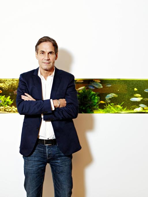 Jean Remy von Matt vor dem Aquarium der Werbeagentur Jung von Matt in der Glashüttenstrasse in Hamburg. Das Aquarium ist in eine sonst komplett weiße Wand eingebettet.