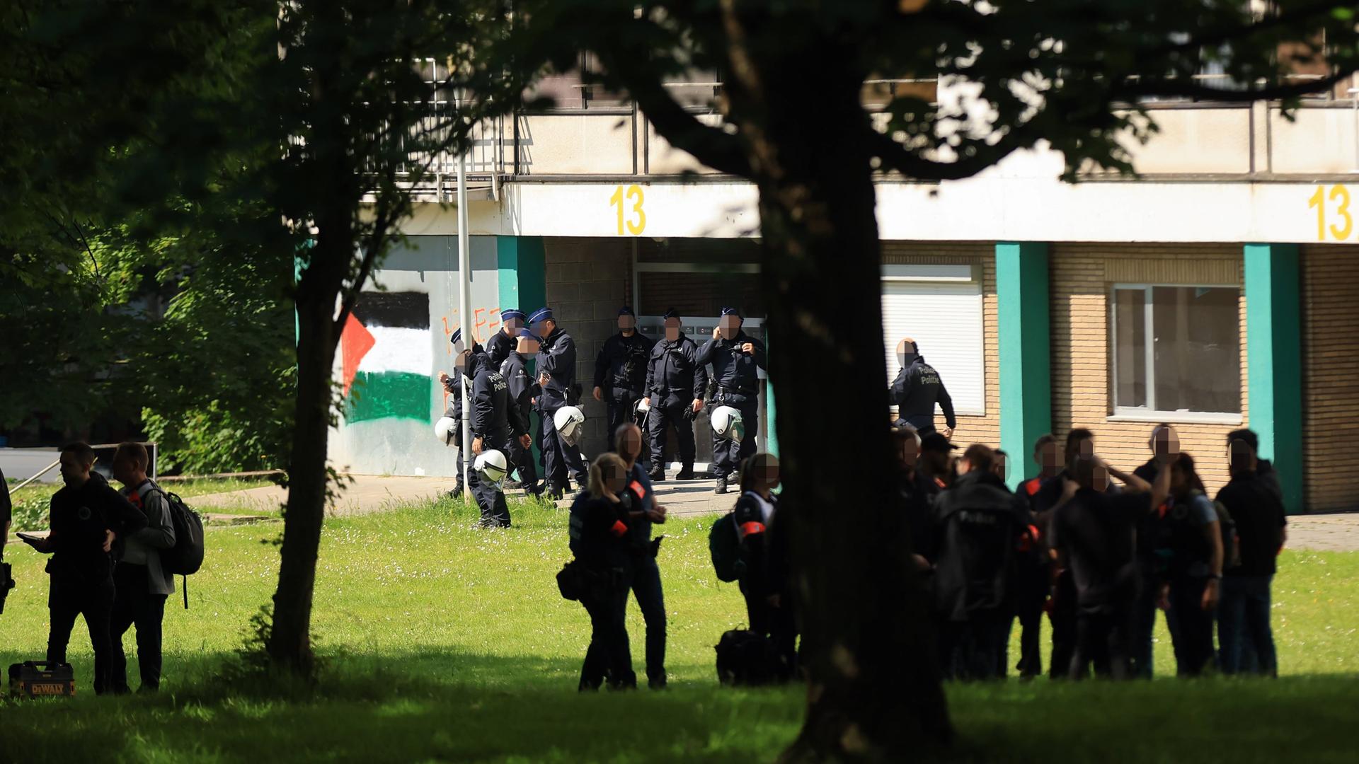 Polizisten riegeln ein Haus in Anderlecht ab und durchsuchen es. Davor stehen Bewohner auf einer Wiese. Polizisten riegeln ein Haus in Anderlecht ab und durchsuchen es. Davor stehen Bewohner auf einer Wiese.