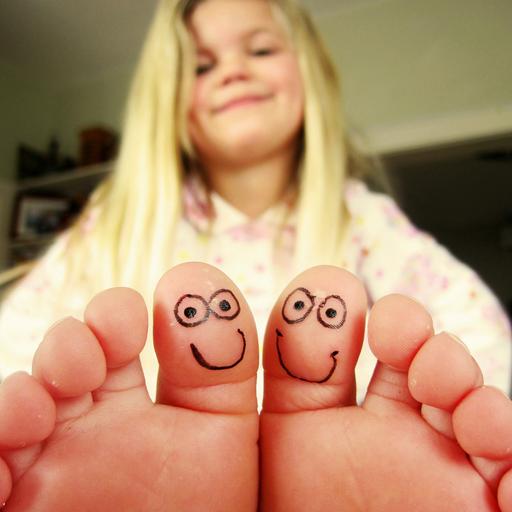 Ein Mädchen sitzt in einem Zimmer und hält ihre Zehen in die Kamera, die beiden großen Zehen sind mit kleinen gesichtern bemalt