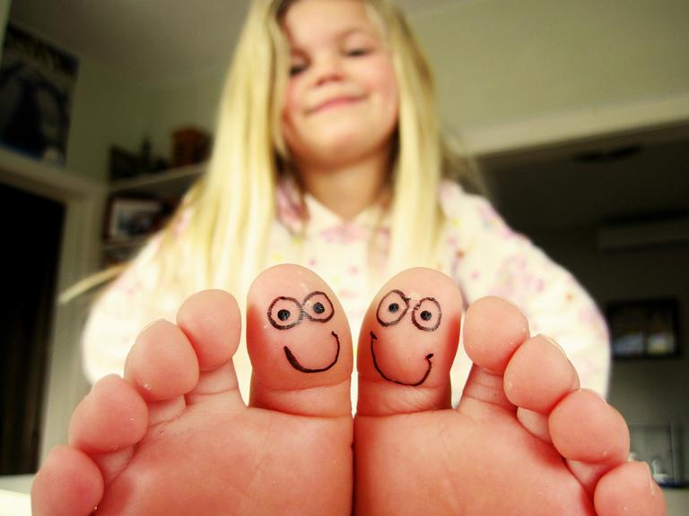 Ein Mädchen sitzt in einem Zimmer und hält ihre Zehen in die Kamera, die beiden großen Zehen sind mit kleinen gesichtern bemalt