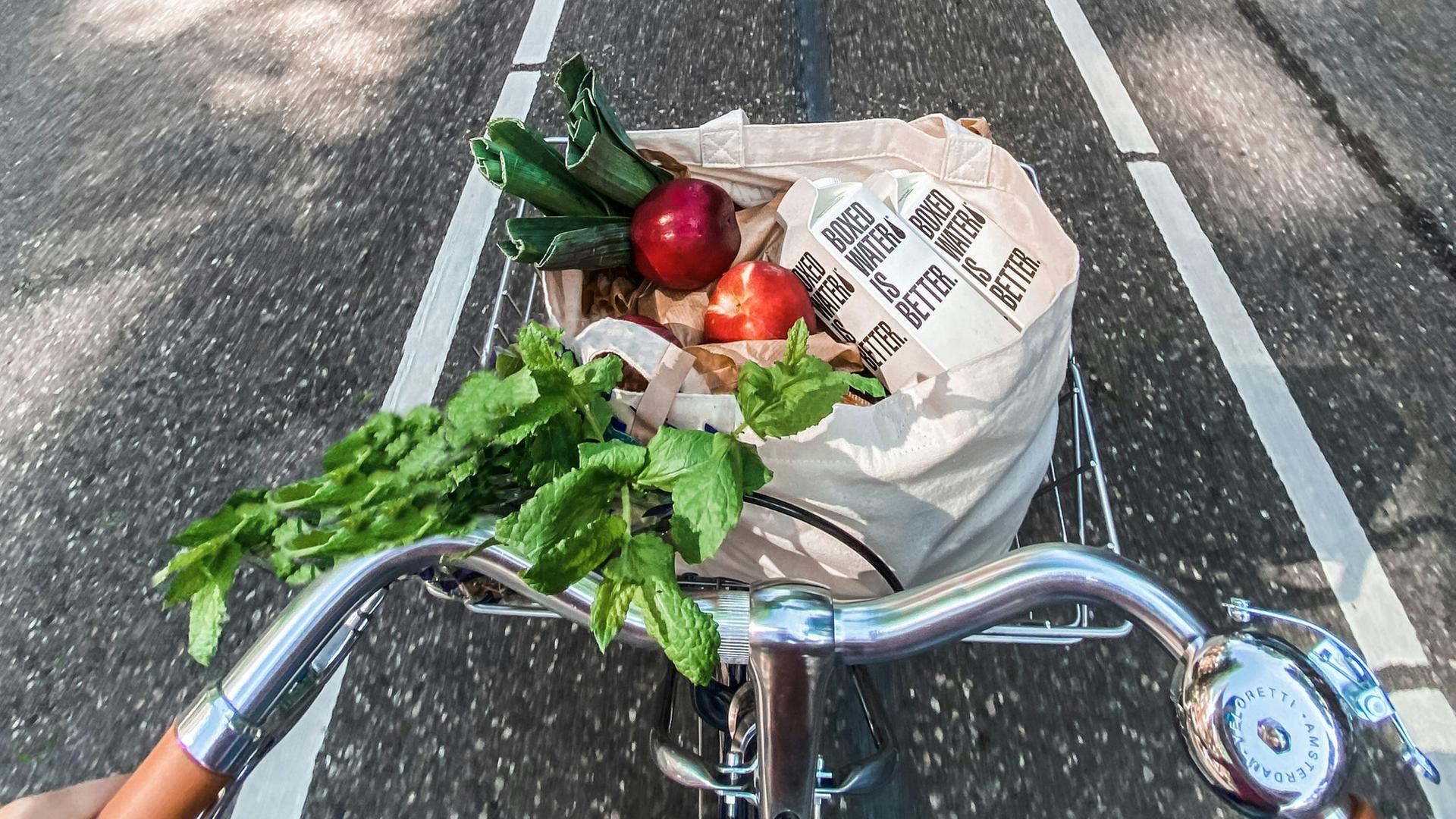 Vogelperspektive auf ein fahrendes Fahrrad, in dessen Korb ein Jutebeutel mit Obst und Gemüse liegt.
