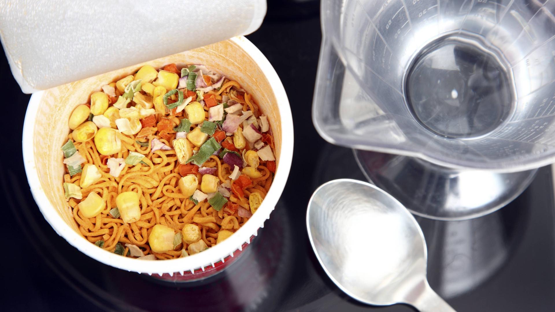 Aufsicht auf eine geöffnete Fertignudelsuppe. Daneben liegen Küchenutensilien. Aufsicht auf eine geöffnete Fertignudelsuppe. Daneben liegen Küchenutensilien.