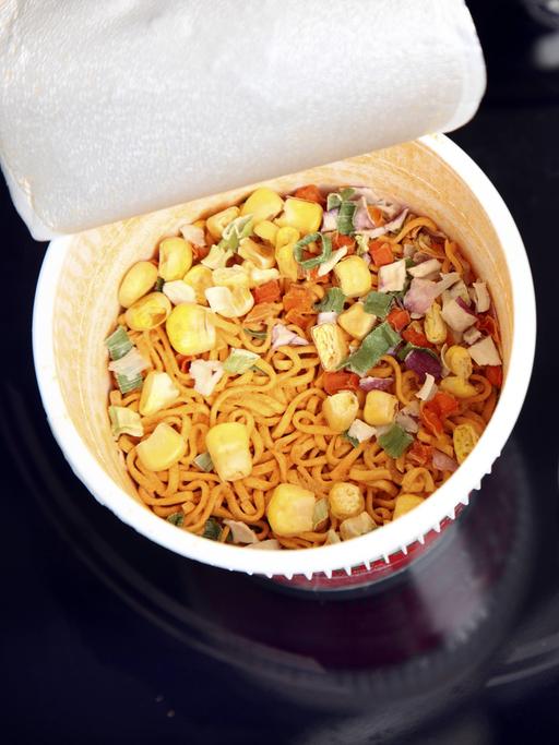 Aufsicht auf eine geöffnete Fertignudelsuppe. Daneben liegen Küchenutensilien.