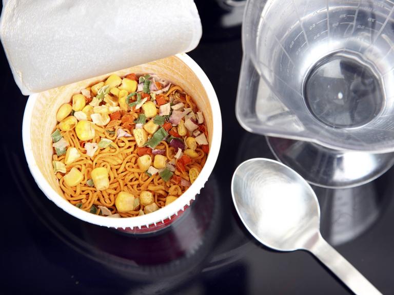 Aufsicht auf eine geöffnete Fertignudelsuppe. Daneben liegen Küchenutensilien.