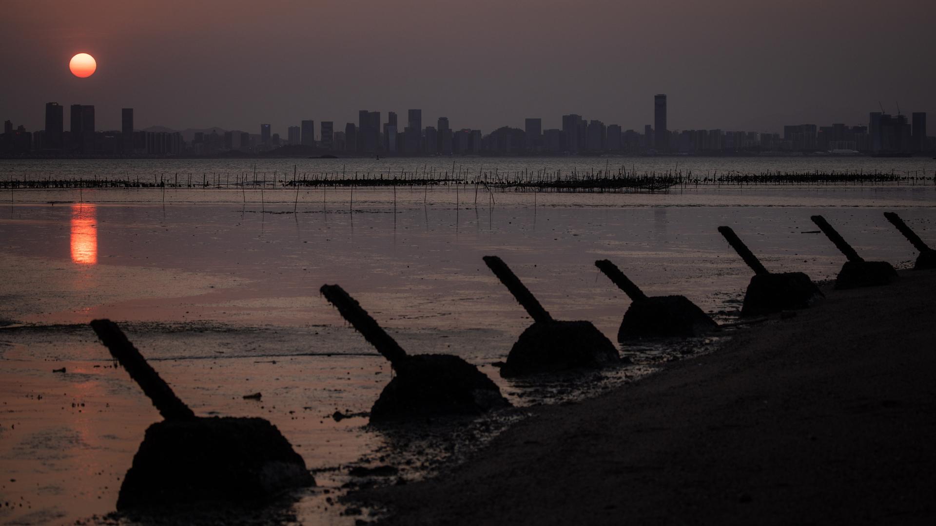 Panzerabwehrbarrikaden stehen am Strand der taiwanischen Insel Kinmen. Im Hintergrund geht über der Silhouette der chinesischen Stadt Xiamen die Sonne unter.