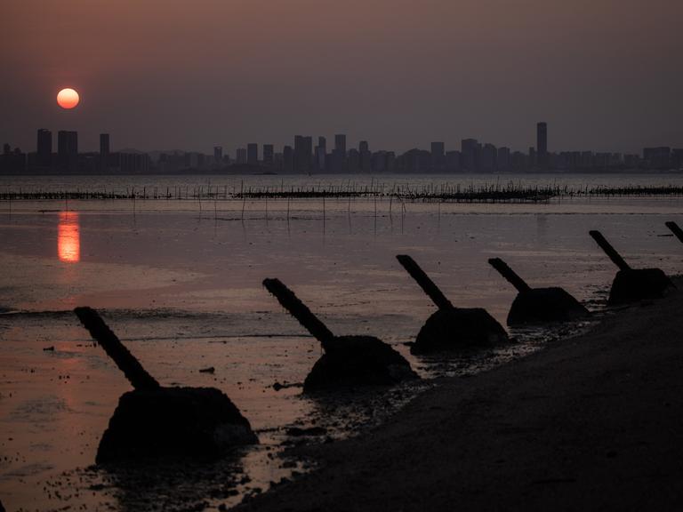 Panzerabwehrbarrikaden stehen am Strand der taiwanischen Insel Kinmen. Im Hintergrund geht über der Silhouette der chinesischen Stadt Xiamen die Sonne unter.