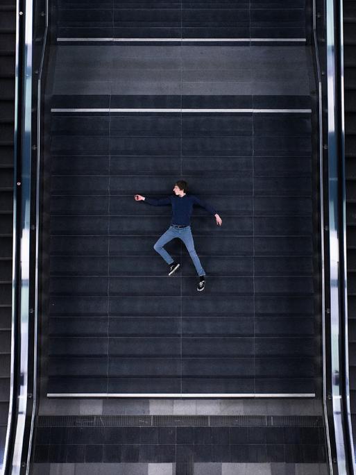 Vogelperspektive auf einen Mann, der zwischen zwei Rolltreppen auf der Treppe liegt.