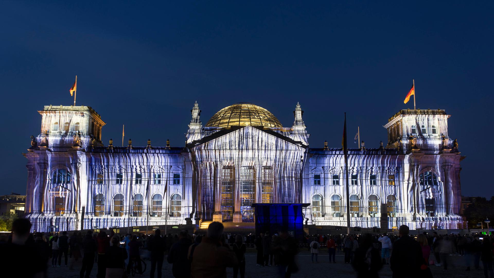 Eine Projektion auf die Westfassade des Reichstags, die aussieht als wäre das Gebäude mit Stoffen behängt. Das Foto wurde bei Nacht aufgenommen. 