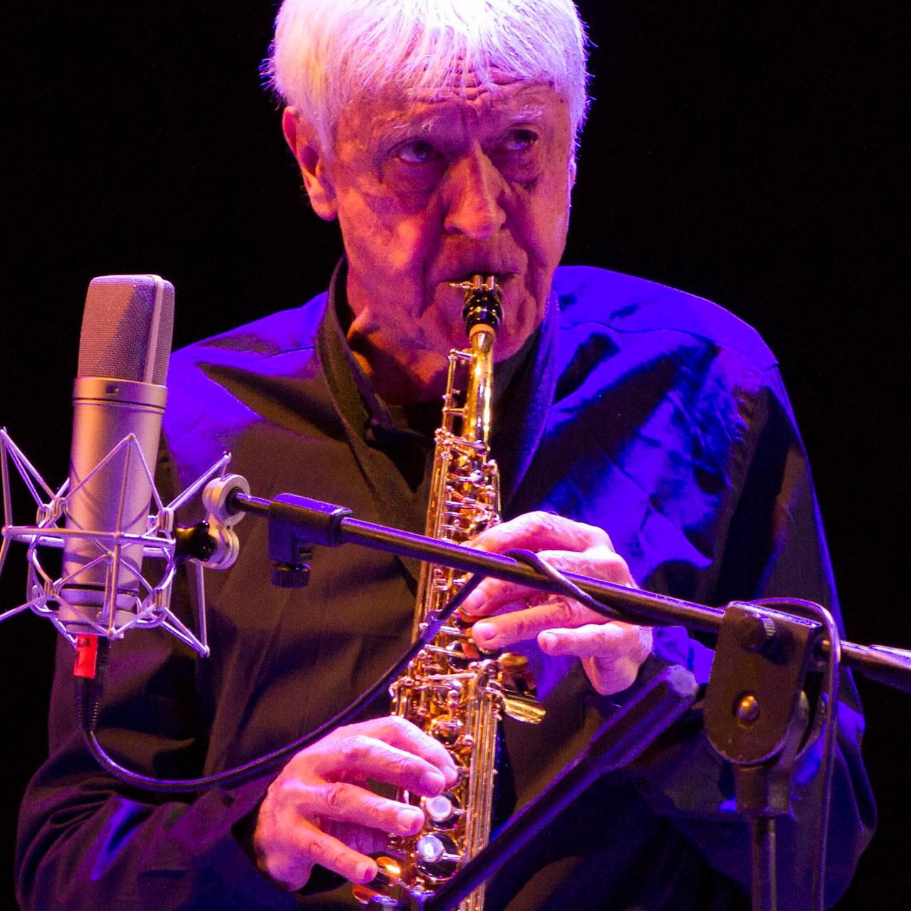 Portal steht auf der Bühn, bläst in sein Instrument und blickt nach vorne. Er wird blau angestraht, der Hintergrund ist dunkel. 